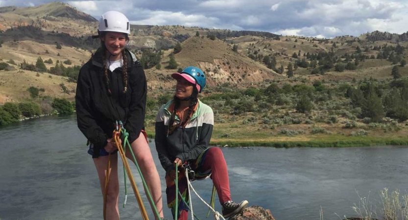 two people wearing rock climbing gear stand on a cliff about water before rappelling down